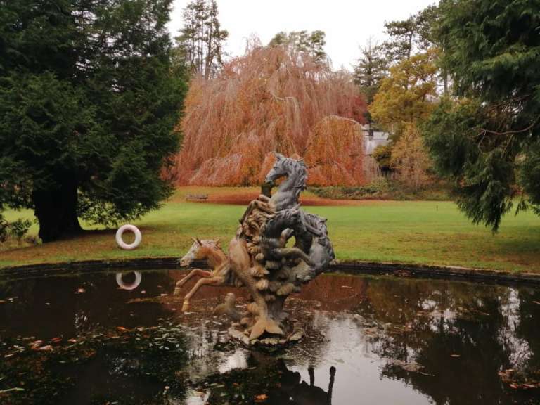 Eine Statue von Pferden in einem Teich vor herbstlich gefärbten Bäumen und einem Gebäude.