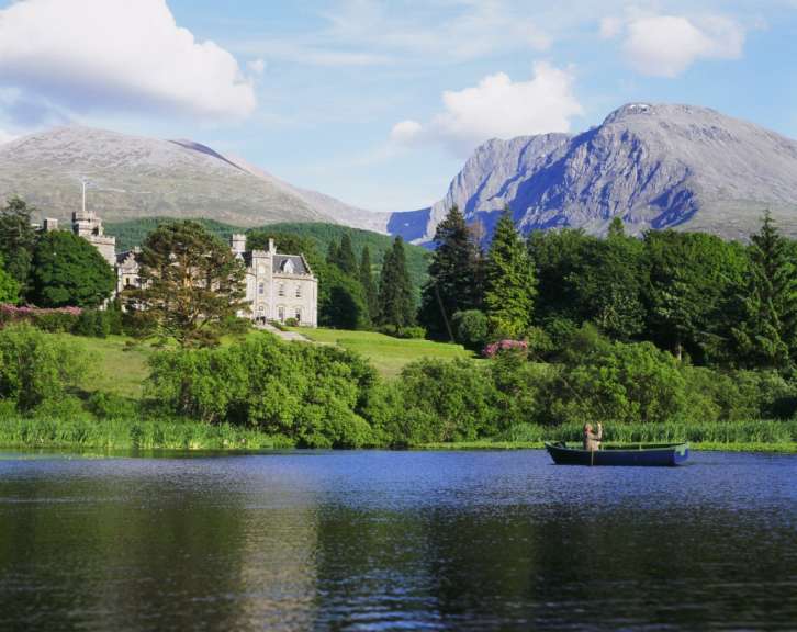 Auf einem See rudert eine Person in einem Boot vor einem Schloss und Bergen.