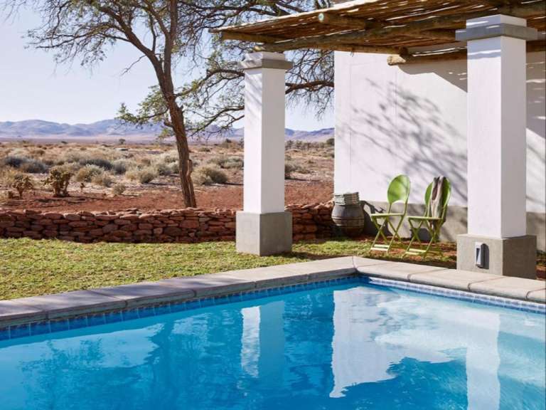Ein Schwimmbecken mit blauem Wasser grenzt an eine grasbewachsene Fläche mit einer Steinmauer und einer Pergola mit zwei grünen Stühlen.