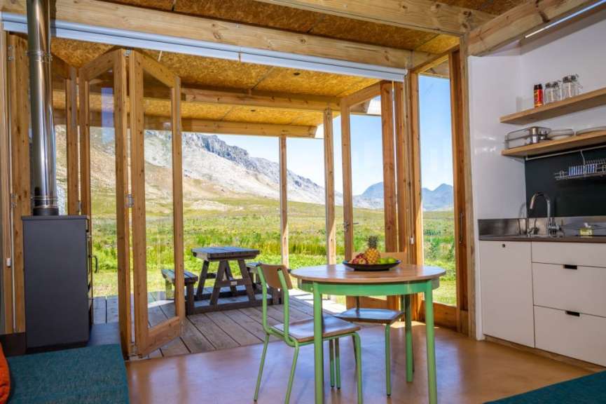 Dieses Bild zeigt ein gemütliches Tiny House mit Holzboden und großen Glasfronten, die einen Blick auf eine Berglandschaft und eine offene Küche freigeben.