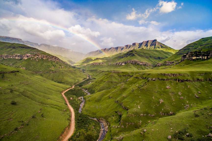 Eine unbefestigte Straße führt durch eine grüne Bergkette mit einem Regenbogen darüber