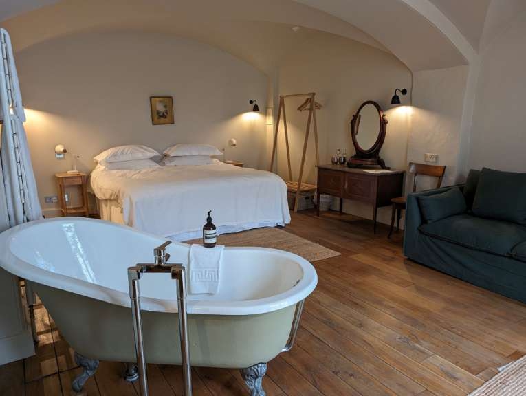 In einem Schlafzimmer mit einer freistehenden Badewanne steht ein Bett, ein Kleiderständer, eine Kommode mit Spiegel und ein Sofa.