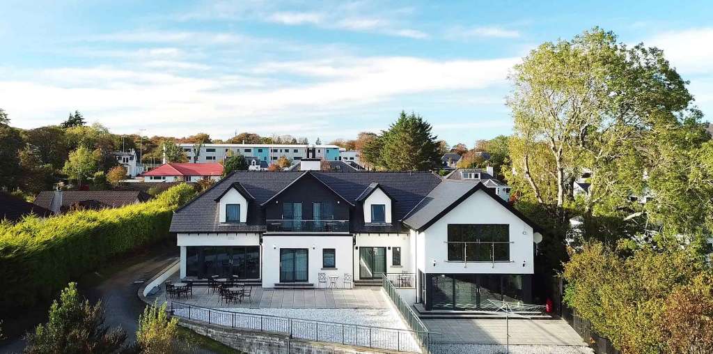 Ein modernes zweistöckiges Haus mit dunklem Dach und weißen Wänden steht auf einem begrünten Grundstück unter einem blauen Himmel mit Wolken.
