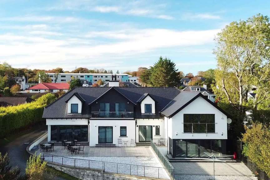 Ein modernes, weißes Haus mit schwarzen Akzenten und großen Fenstern, das von Bäumen und Büschen umgeben ist, steht auf einem terrassierten Grundstück.