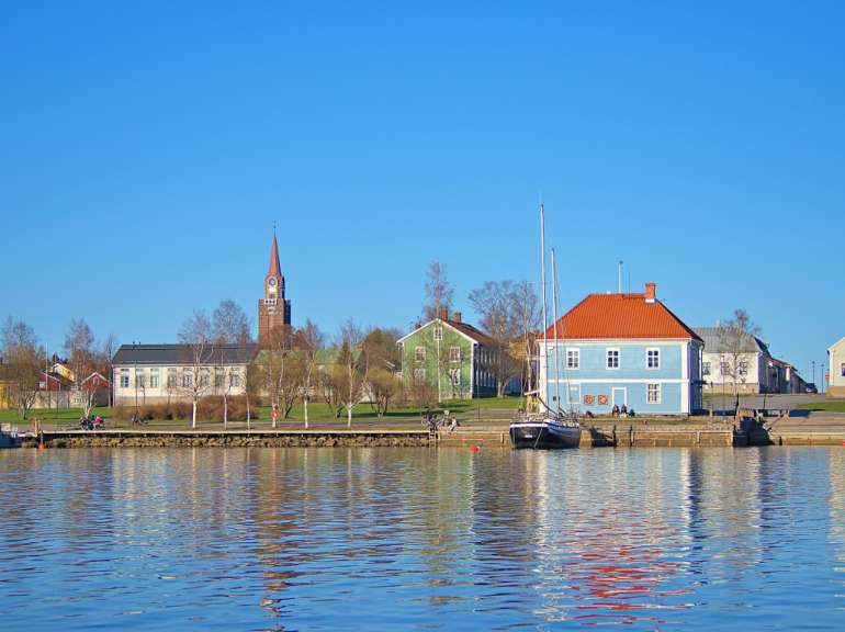 Ein blauhäusiges Segelboot liegt im Hafen vor einer Reihe von Gebäuden, darunter ein kirchturmähnliches Gebäude und ein grünes und ein weiteres blaues Haus.