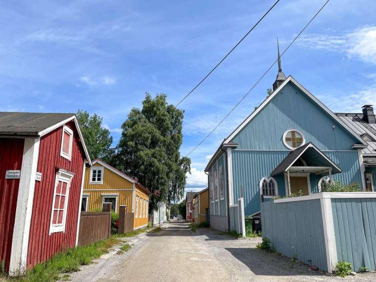 Auf dem Bild ist eine sonnige Gasse mit bunten Holzhäusern und einer blauen Kirche zu sehen.