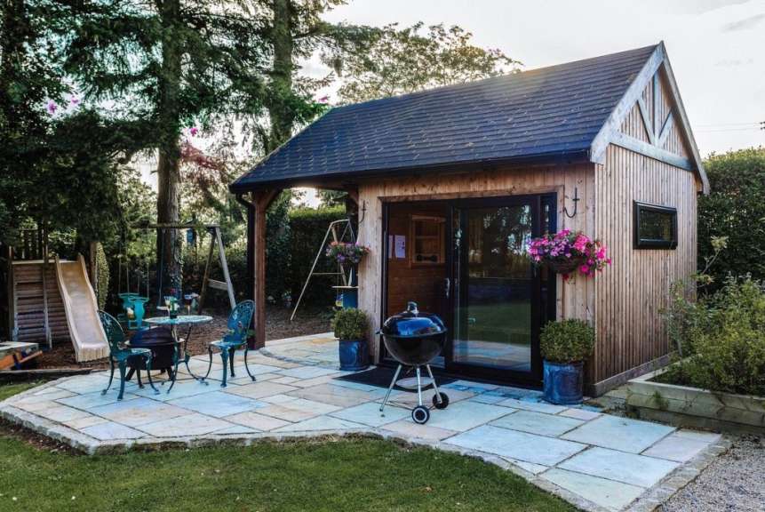 Ein Holzgartenhaus mit Veranda und zwei Stühlen sowie einem Grill steht auf einer Steinterrasse vor einem Rasen und Bäumen.