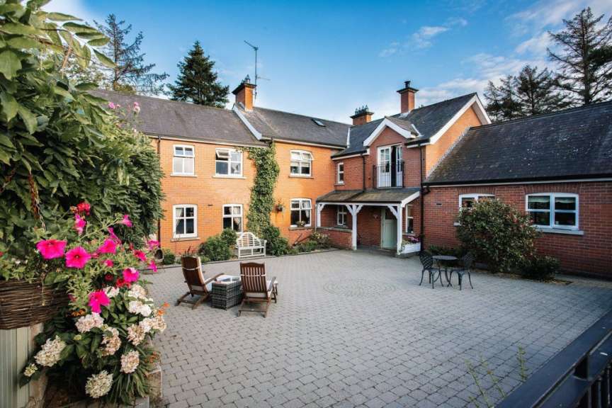 Ein Ziegelhaus mit einem gepflasterten Hof und einem Sitzbereich im Freien wird von Bäumen und Blumen umgeben.