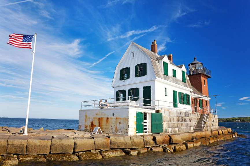 Ein Leuchtturm mit einem weißen Haus und einer amerikanischen Flagge auf einem Felsvorsprung im Meer unter einem blauen Himmel.