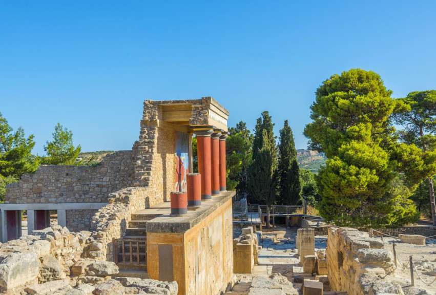 Die Ruinen des Palastes von Knossos auf Kreta werden von roten Säulen und einer Wandmalerei einer Stierszene geschmückt, während Bäume und eine hügelige Landschaft im Hintergrund zu sehen sind.