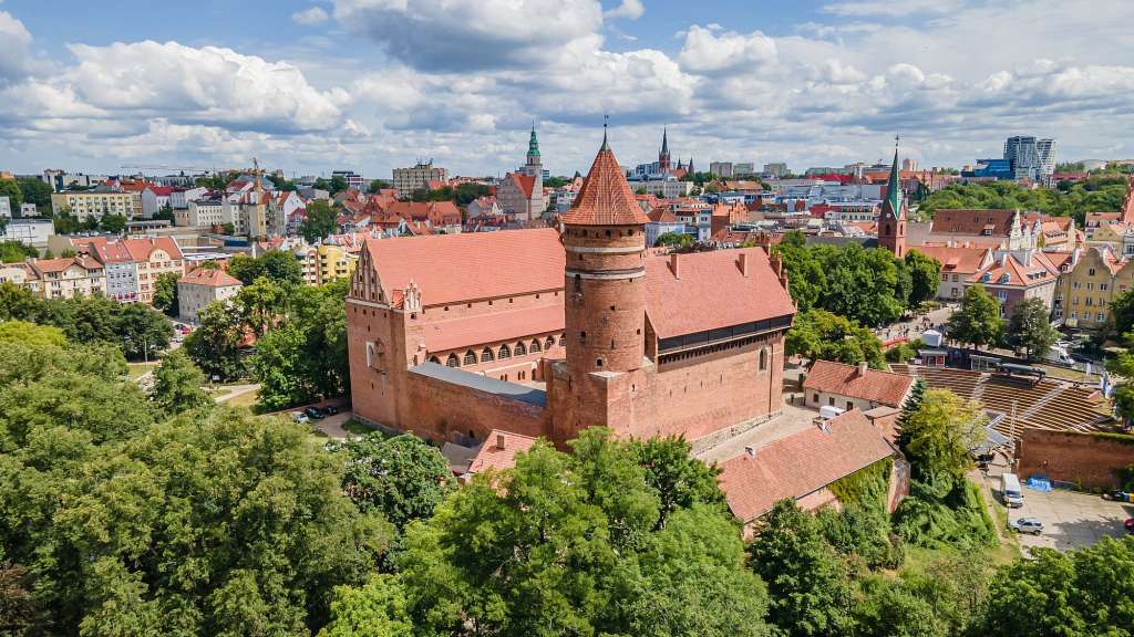 Die Luftaufnahme zeigt eine mittelalterliche Backsteinburg in einer Stadt mit roten Dächern und viel Grün.