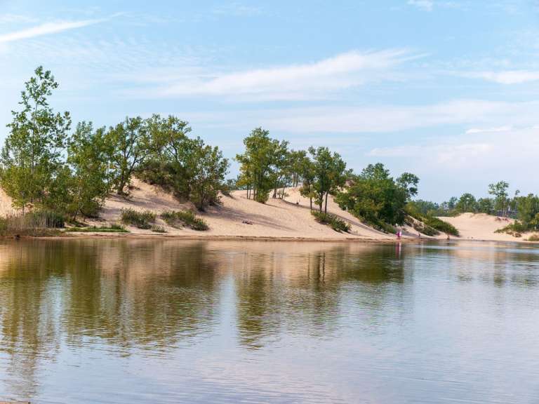 Eine Person steht am Ufer eines Sees mit Bäumen und Sanddünen im Hintergrund