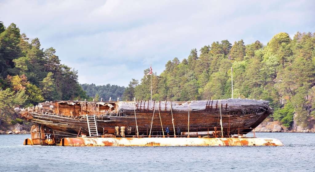 Ein verlassenes Holzschiff schwimmt im Wasser, umgeben von Bäumen und bewölktem Himmel.