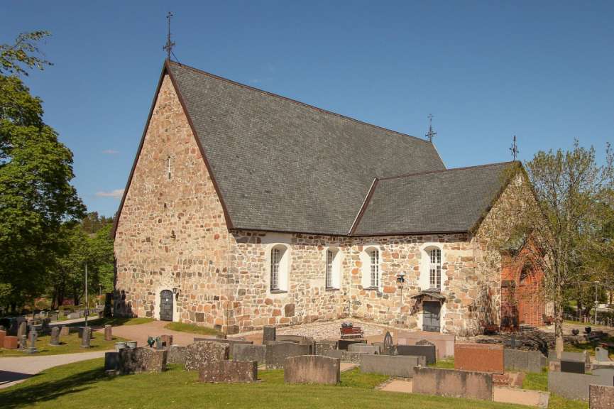 Eine Steinkirche mit Ziegelfassade steht auf einem Friedhof mit vielen Grabsteinen unter einem blauen Himmel.