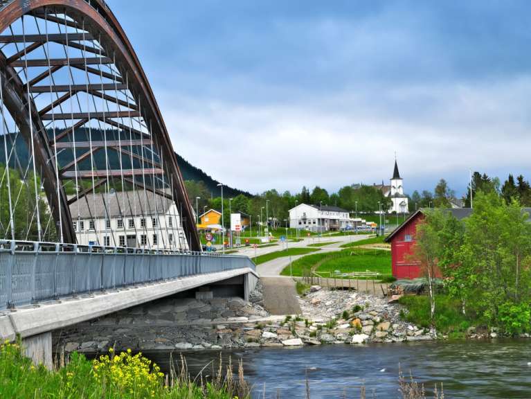 Eine Brücke überspannt einen Fluss vor einer ländlichen Kulisse mit Häusern und einem Kirchturm.