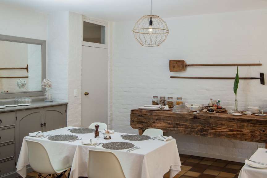 Ein Esstisch mit weißer Tischdecke ist in einem hellen Raum gedeckt, daneben steht ein Buffet mit Gläsern und Lebensmitteln.