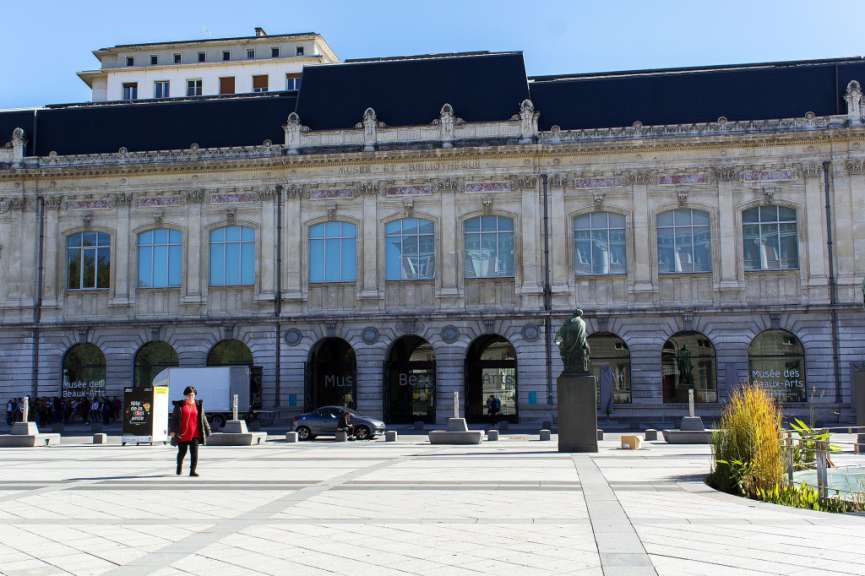 Eine Frau geht durch eine sonnige, gepflasterte Straße vor einem großen Gebäude mit Bogengängen und Fenstern, die eine breite Steinfassade und den blauen Himmel widerspiegeln.