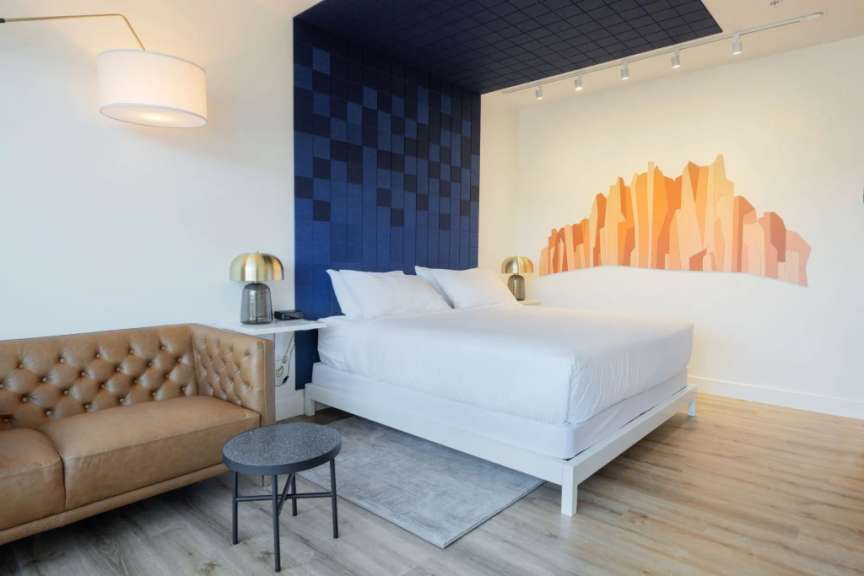 Ein modernes Schlafzimmer mit einem beigen Ledersofa, einem weißen Bett, zwei Nachttischen mit Lampen und einer Wanddekoration in Form einer bergähnlichen Silhouette.