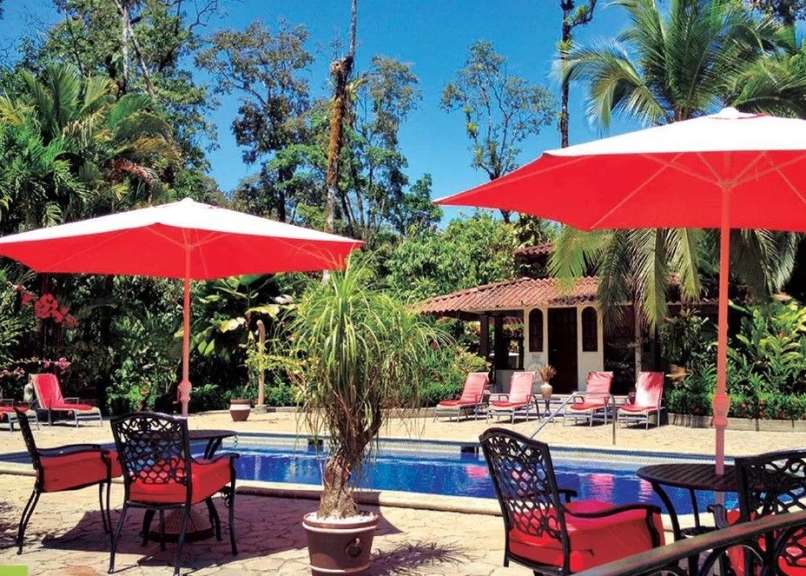 Ein tropischer Innenhof mit Pool, umgeben von üppiger Vegetation und mit roten Sonnenschirmen, Liegestühlen und Tischen.