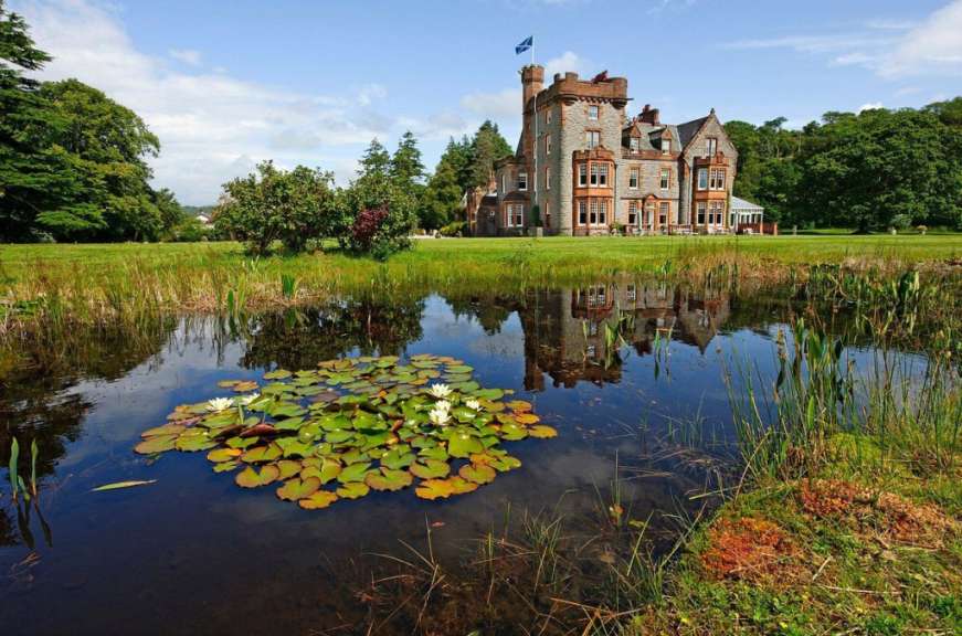 Ein Schloss im schottischen Stil mit einem Teich und Seerosen davor spiegelt sich im Wasser an einem sonnigen Tag.