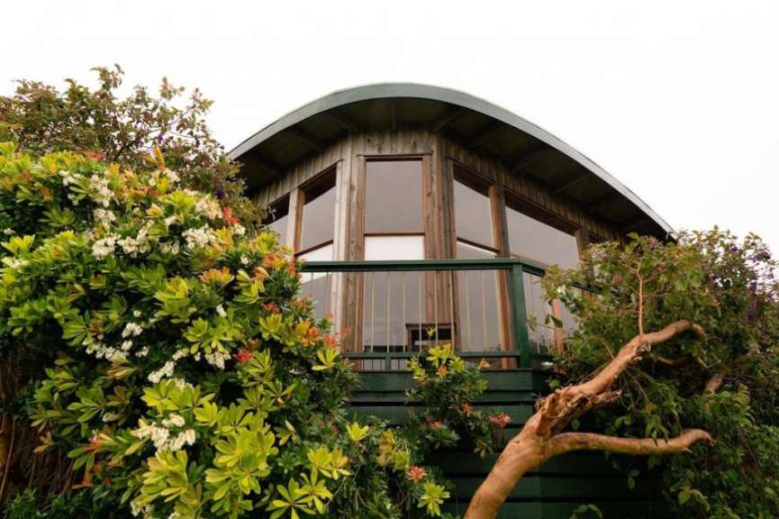 Ein zweistöckiges Holzhaus mit gebogenem Dach und großen Fenstern, das von üppiger Vegetation und Bäumen umgeben ist.