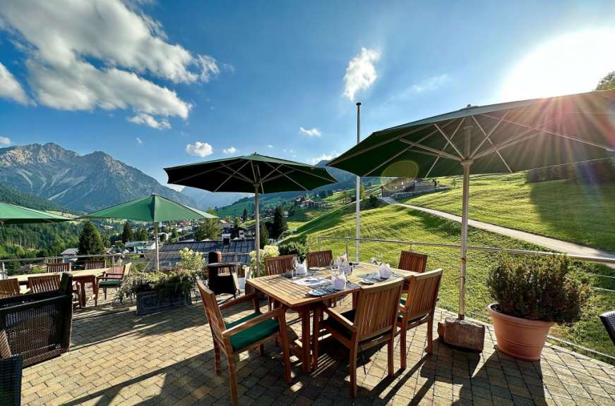 Ein gedeckter Tisch mit Stühlen steht auf einer Terrasse mit Berg- und Wiesenblick unter Sonnenschirmen.