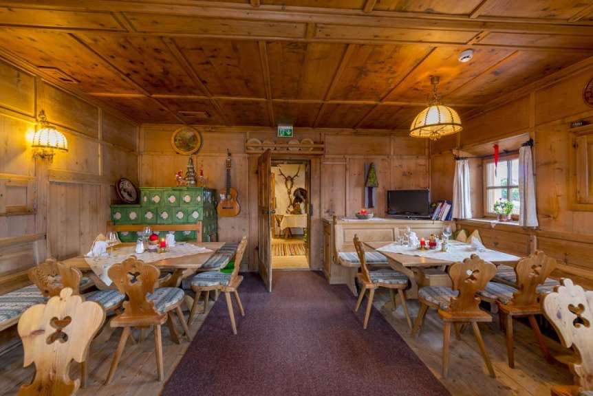 In einem holzgetäfelten Speisesaal mit traditionellen Möbeln stehen gedeckte Tische mit Kerzen, ein grüner Kachelofen, eine Gitarre und ein Fernseher.