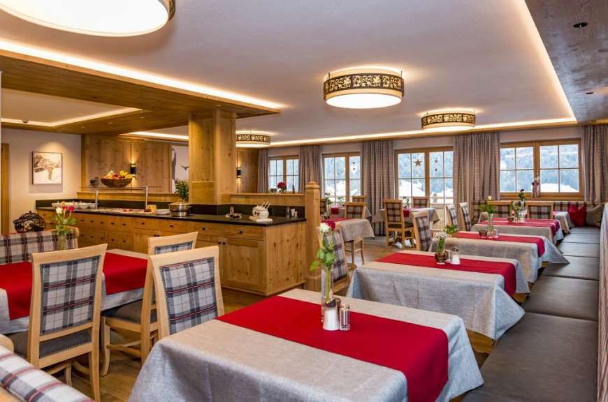 In einem rustikalen Speisesaal sind Tische mit roten Tischläufern gedeckt und von Holzsitzbänken und karierten Stühlen umgeben, während im Hintergrund ein Buffetbereich zu sehen ist.