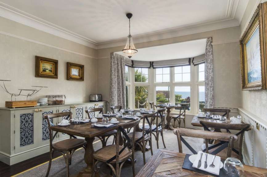 Ein Essbereich mit Tischen und Stühlen, die mit Frühstücksgeschirr für eine Mahlzeit gedeckt sind, steht im Hintergrund eines fenstergesäumten Raumes mit Blick auf das Meer.