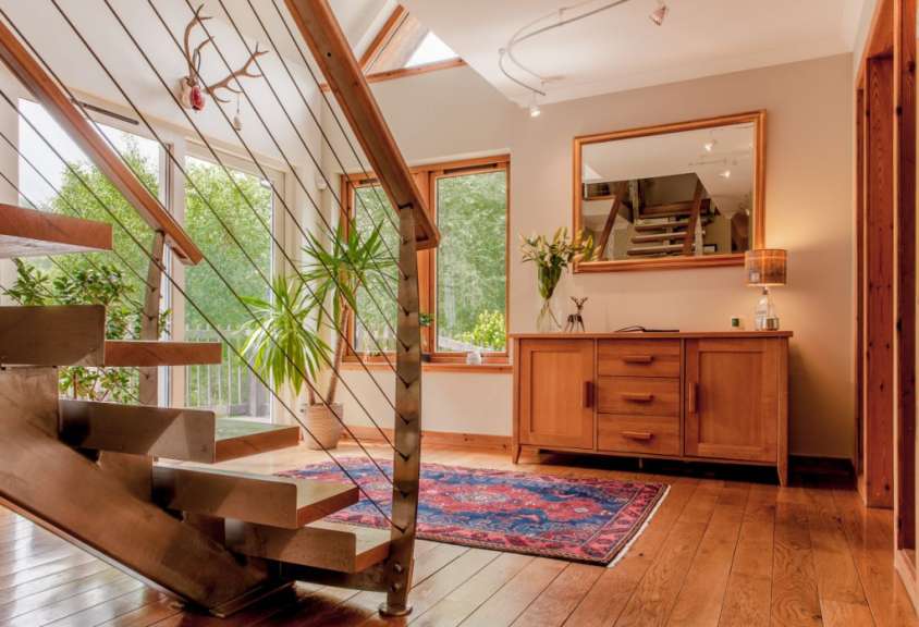 In einem modern eingerichteten Haus mit Holztreppe und Spiegel steht eine Anrichte mit einem Blumenstrauß und einer Lampe vor einem Fenster.