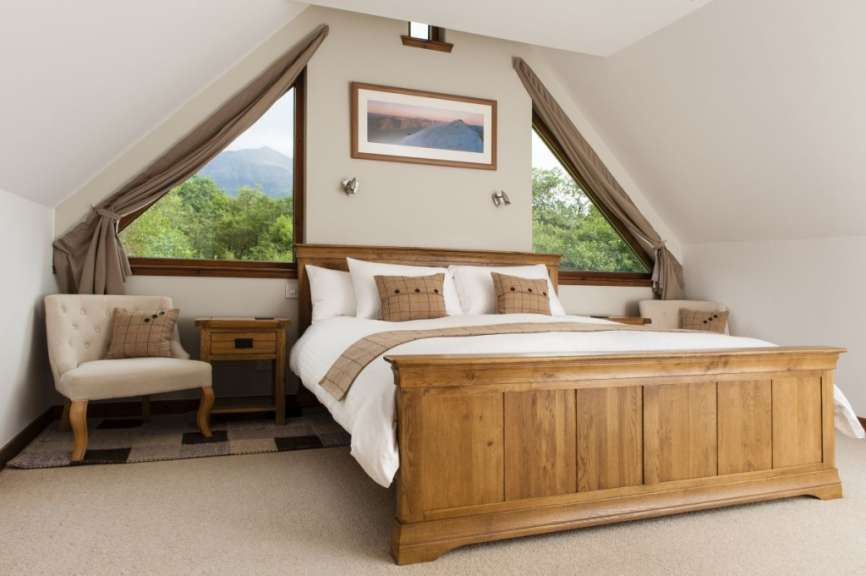 Ein großes Himmelbett aus Holz steht in einem gemütlichen Schlafzimmer mit Dachschrägenfenstern, die auf einen Wald und Berge blicken.