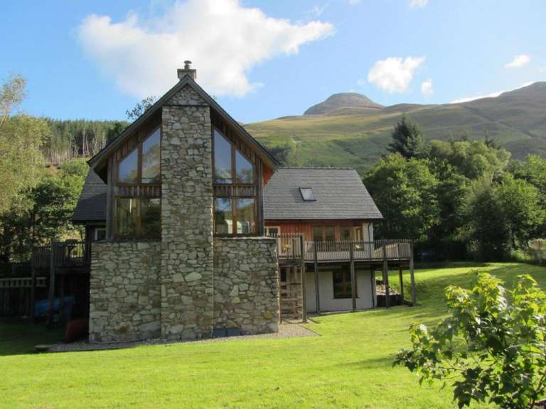 Ein zweigeschossiges Haus aus Stein und Holz mit großen Fenstern und einem Holzdeck steht auf einer grünen Wiese vor einem bewaldeten Hügel und einem Berg unter blauem Himmel mit Wolken.