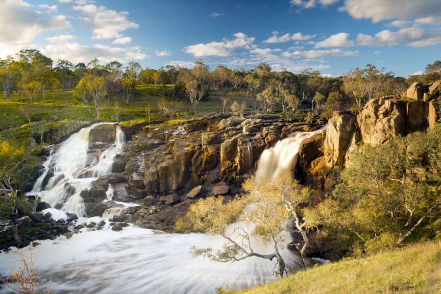Ein malerischer Wasserfall fließt über felsige Klippen in einer baumgesäumten Landschaft unter einem strahlenden Himmel mit Wolken.