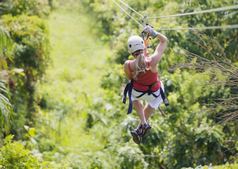 Eine Frau in einem roten Tanktop und weißen Shorts, die einen Helm trägt, seilrutscht über einen üppigen grünen Dschungel.