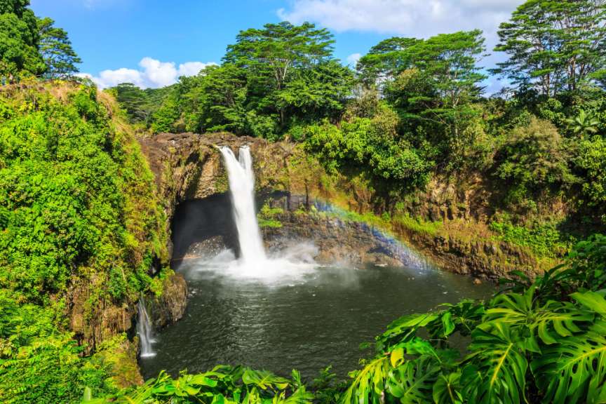 Ein Regenbogen bildet sich in der Gischt eines Wasserfalls, der in ein türkisfarbenes Wasserbecken stürzt, umgeben von üppiger grüner Vegetation unter einem blauen Himmel mit weißen Wolken.