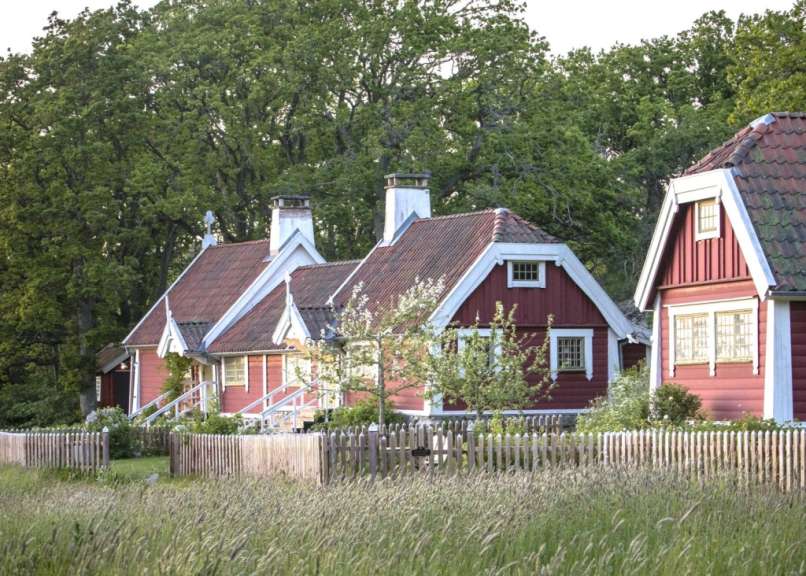 Auf diesem Bild sind drei rote Schwedenhäuser mit weißen Fensterrahmen und roten Ziegeldächern von einem Holzzaun im Vordergrund verdeckt.