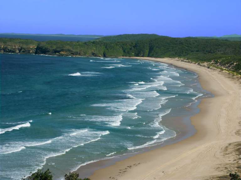 Ein ruhiger Strand mit grünen Hügeln im Hintergrund und aufgewühlten Wellen im Vordergrund.