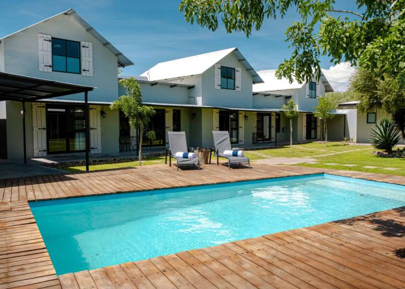 Ein Swimmingpool mit Holzdeck und Liegestühlen vor einer Reihe von kleinen Häusern mit weißen Fensterläden und Metallrohrdächern unter blauem Himmel.