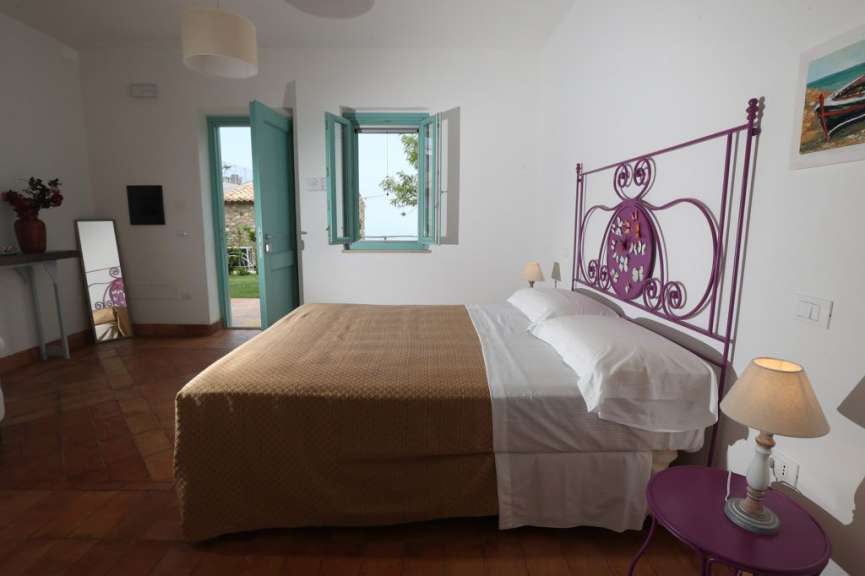 Ein Schlafzimmer mit einem großen Bett, einem lila-farbenen Kopfteil und einem geöffneten Fenster.