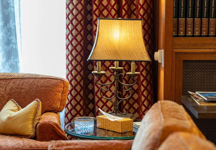 Ein Tisch mit einer Lampe, einem Taschentücher-Spender und einem Aschenbecher steht neben einem Sessel vor einem gemusterten Vorhang.