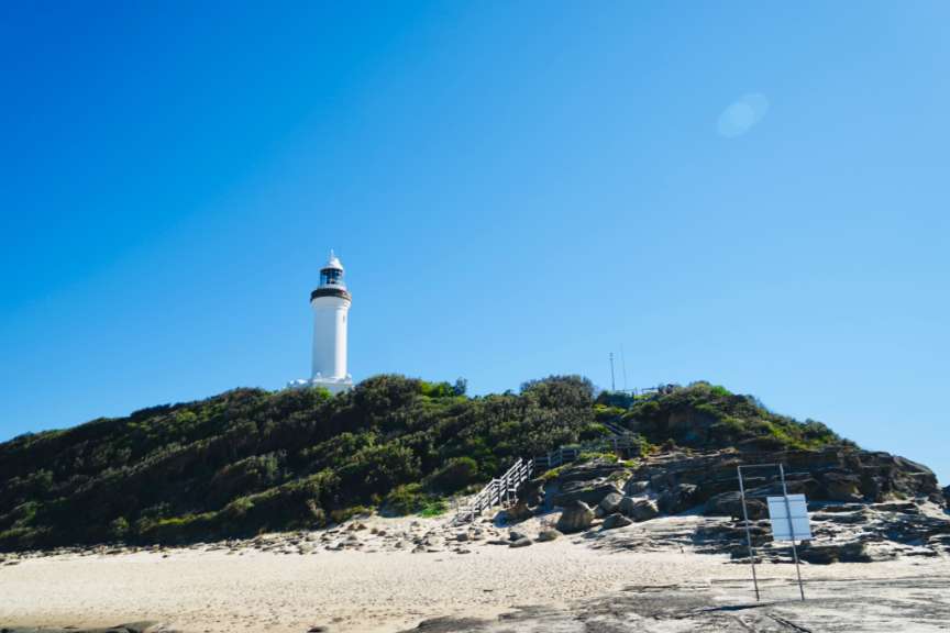 Ein weißer Leuchtturm steht auf einem bewachsenen Hügel über einem sandigen Strand und Felsen unter einem blauen Himmel.