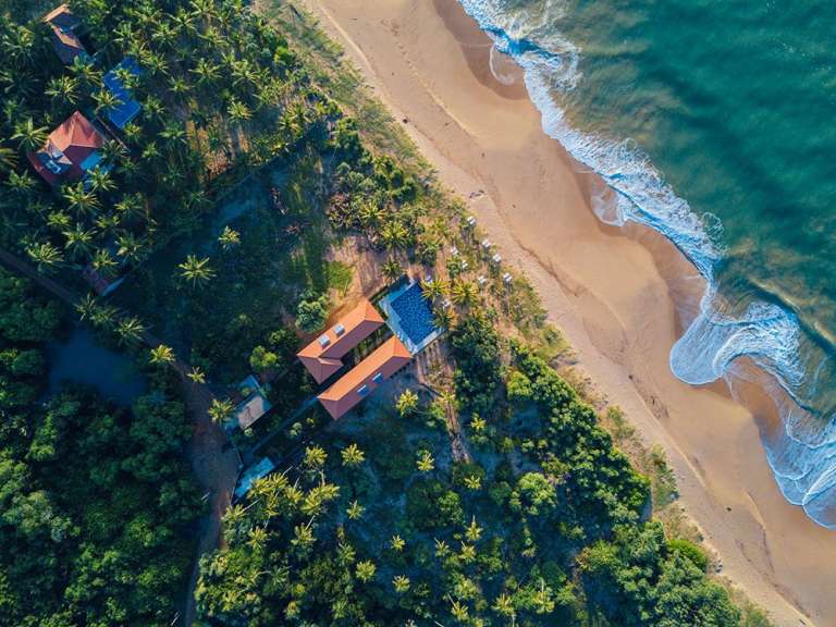 Diese Luftaufnahme zeigt einen tropischen Strand mit Wellen, der von Palmen und einem Resort gesäumt wird.