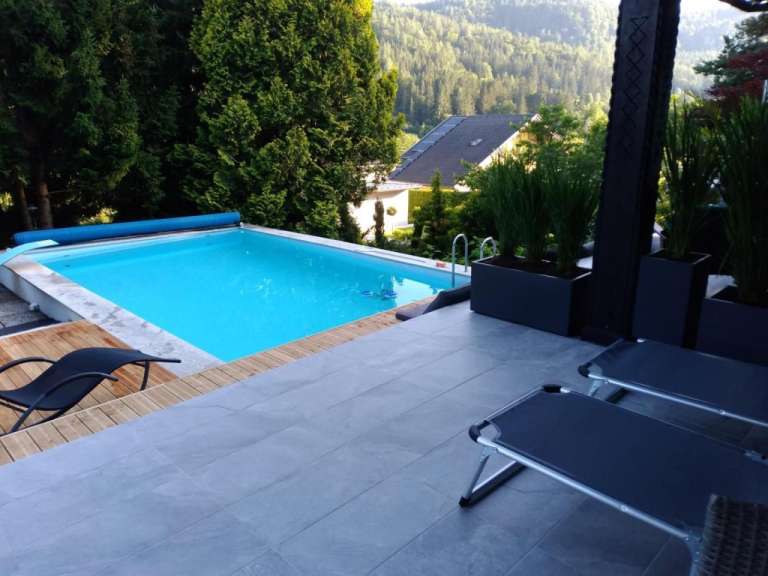 Ein Schwimmbecken mit blau abgedeckter Plane steht neben einem Holzdeck mit Liegestuhl, und zwei weitere Liegestühle stehen auf einer grauen gefliesten Terrasse, während im Hintergrund ein Haus und ein bewaldeter Hang zu sehen sind.