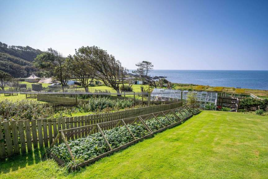 Ein weitläufiger Garten mit Gemüsebeeten, Gewächshäusern und einer Zeltstruktur im Hintergrund blickt auf das Meer.