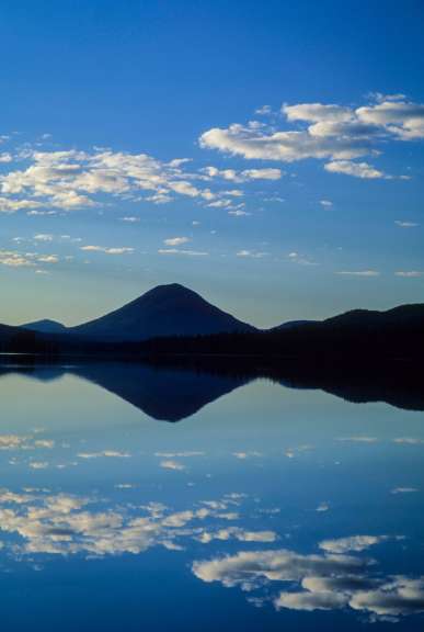 Ein symmetrisches Bild zeigt eine Berglandschaft, die sich im ruhigen Wasser eines Sees spiegelt, unter einem blauen Himmel mit weißen Wolken.