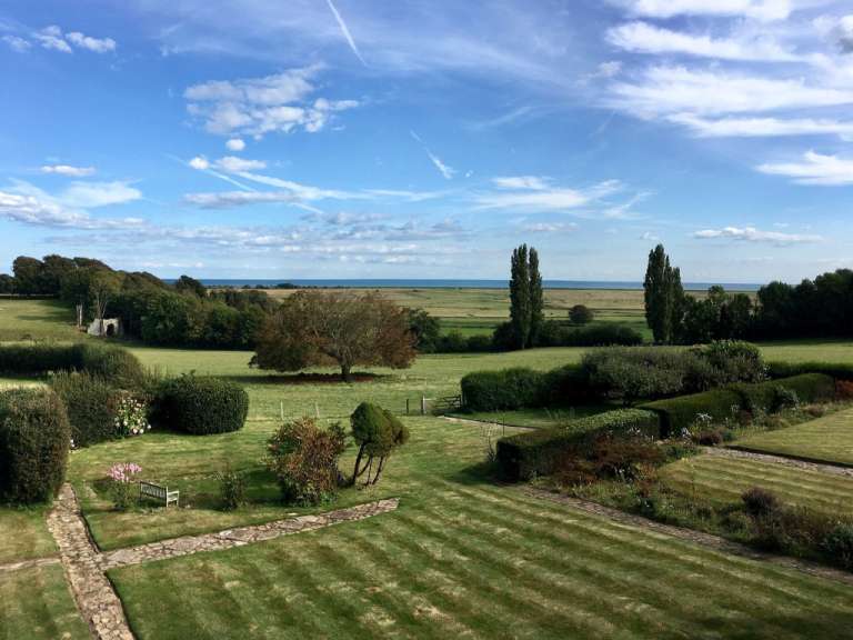 Ein weiter Blick über eine grüne Landschaft mit Bäumen, Büschen und einem gestreiften Rasen unter einem blauen Himmel mit Wolken.