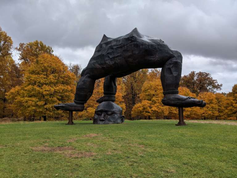 Eine dunkle Skulptur eines riesigen Körpers, der auf einem Kopf ruht und mit den Füßen auf Sockeln steht, ist vor einem Hintergrund mit leuchtend gelben Bäumen zu sehen.