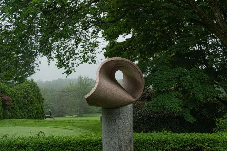Ein grauer Steinmetzkunstwerk steht auf einem Sockel vor einem bewaldeten Hügel und einer bronzefarbenen Skulptur eines Pferdes auf einem Rasen.