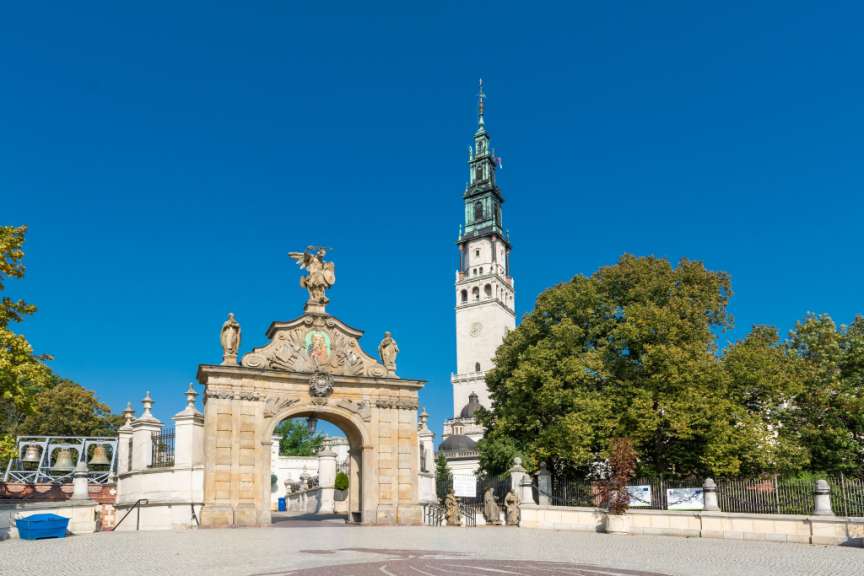 Ein hoher Kirchturm ragt über einer Toranlage und Bäumen vor einem strahlend blauen Himmel auf.