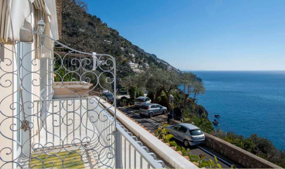 Ein Blick von einem Balkon auf eine kurvige Straße mit Autos, die am Hang eines felsigen Küstenortes mit Blick auf das blaue Meer liegen.